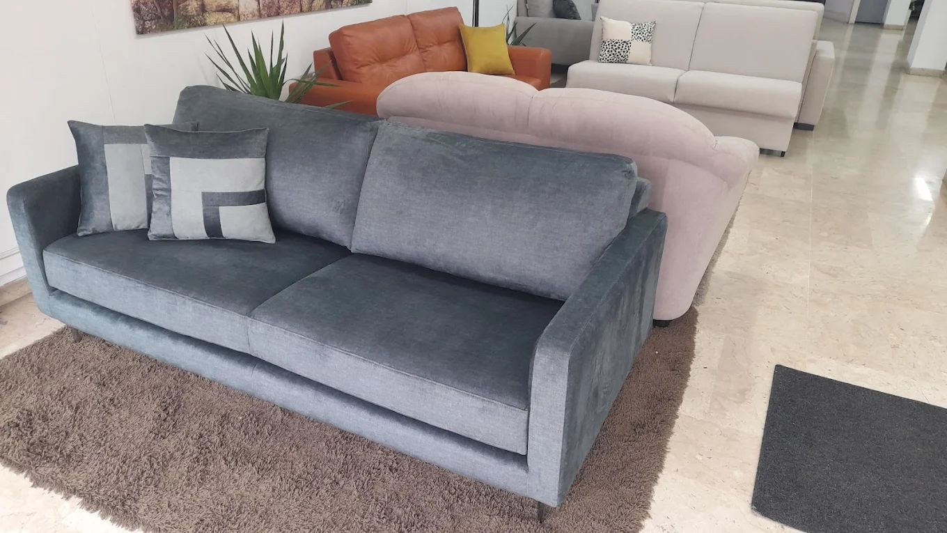 Divano in velluto grigio con cuscini decorativi su un tappeto marrone in uno showroom.