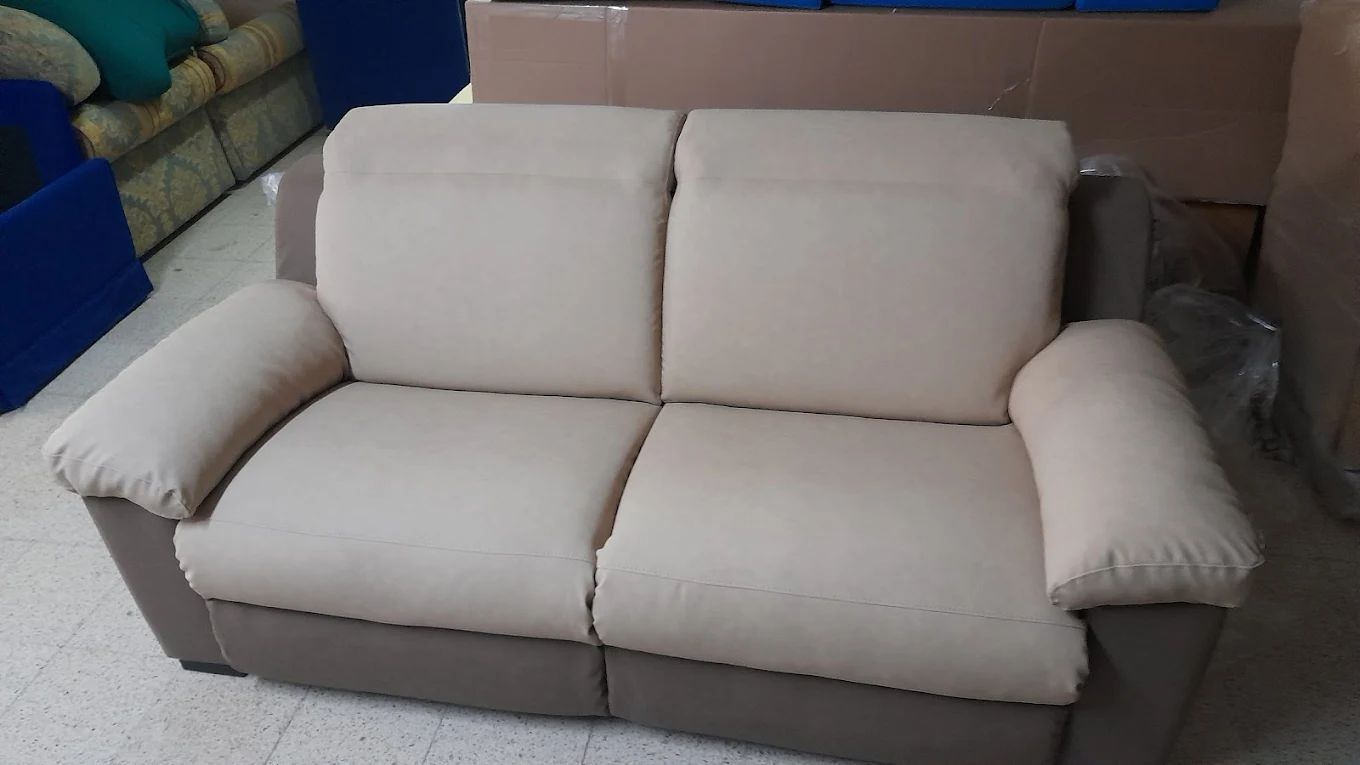 Divano a due posti beige e grigio con schienale imbottito e braccioli arrotolati in un ambiente da negozio.