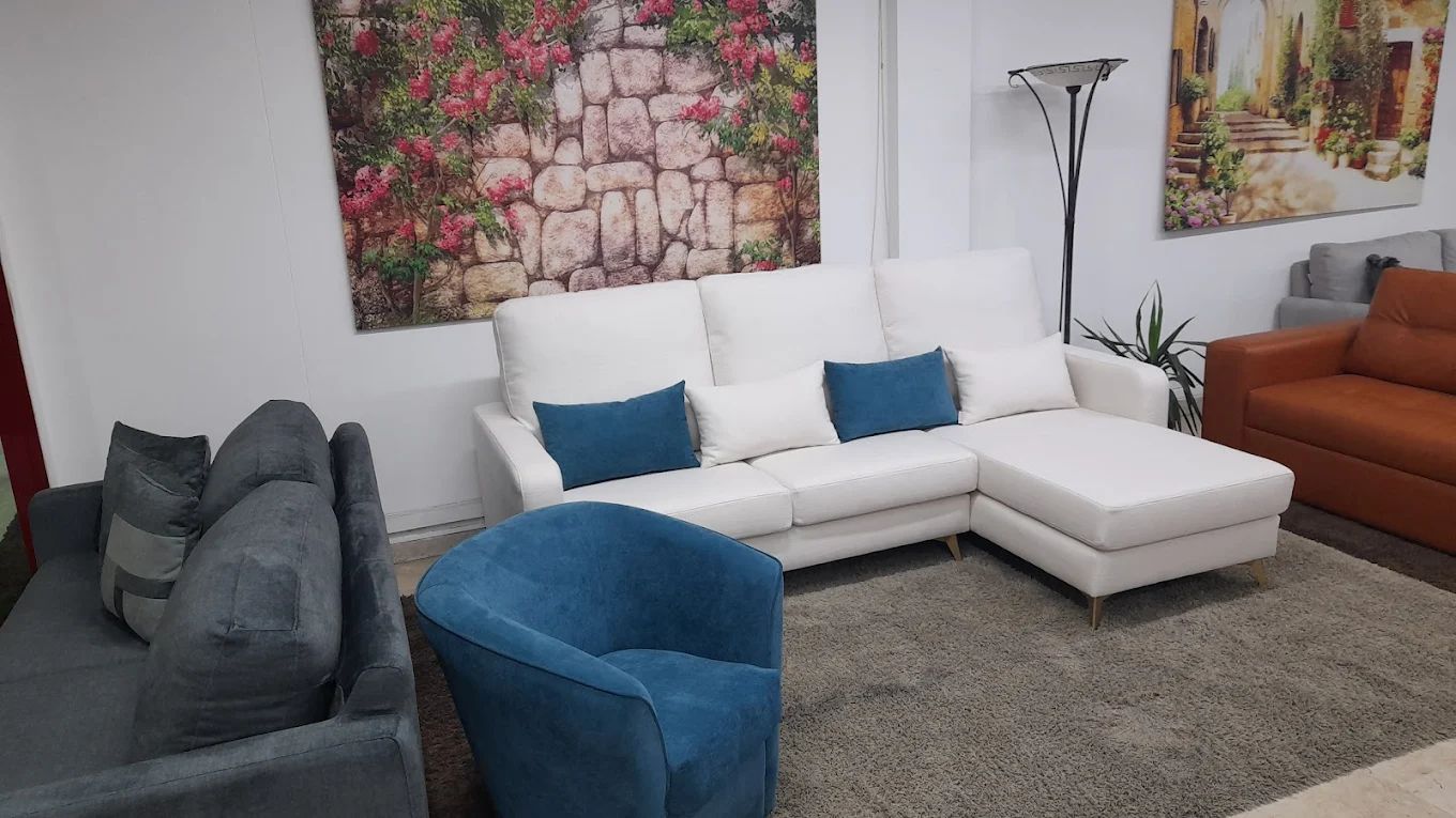 Divano componibile bianco con cuscini blu, sedia blu e tappeto grigio in uno showroom.