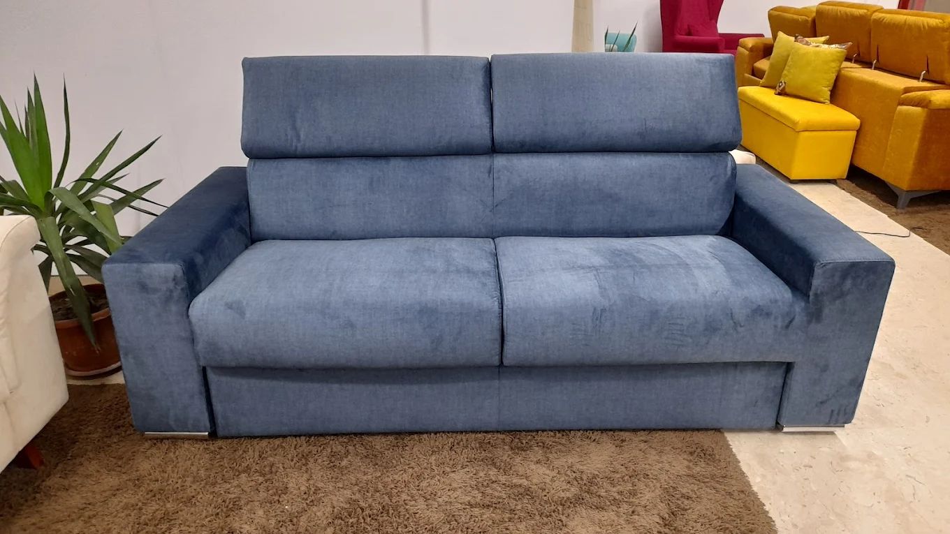 Divano blu in uno showroom, con una pianta sulla sinistra e altri mobili sullo sfondo.