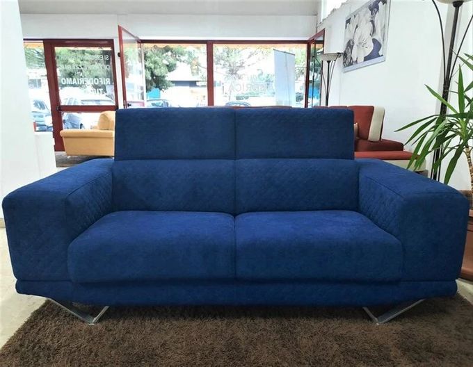 Divano blu a due posti con schienale alto e gambe argentate, esposto in uno showroom.