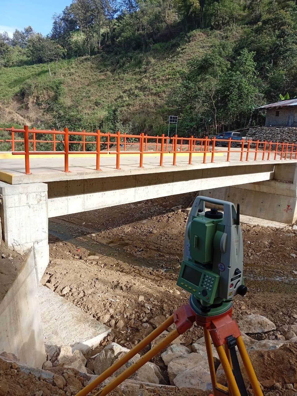 Equipo topográfico instalado cerca de un puente de hormigón con barandillas de color naranja, en un entorno rural.
