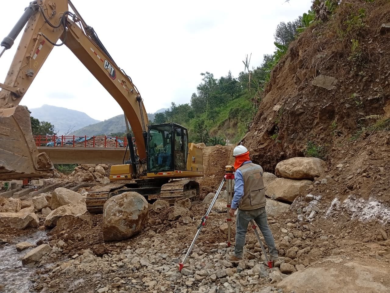 Trabajador de la construcción con equipo topográfico cerca de excavadora y escombros rocosos, puente al fondo.