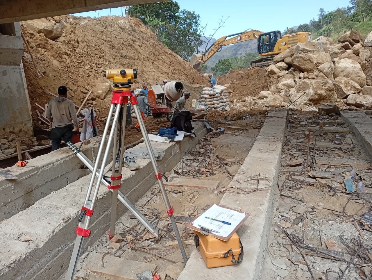 Sitio de construcción con equipo de topografía y trabajadores; excavadora al fondo.