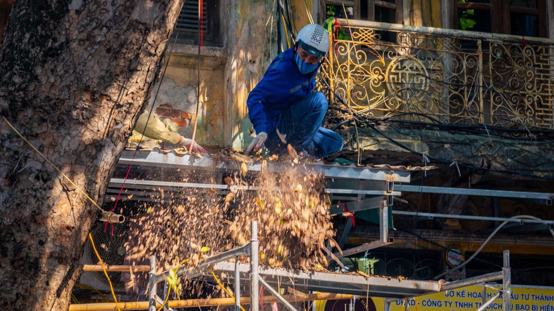 Trabajador de la construcción con mono azul cortando una rama, mientras los escombros caen sobre un andamio.