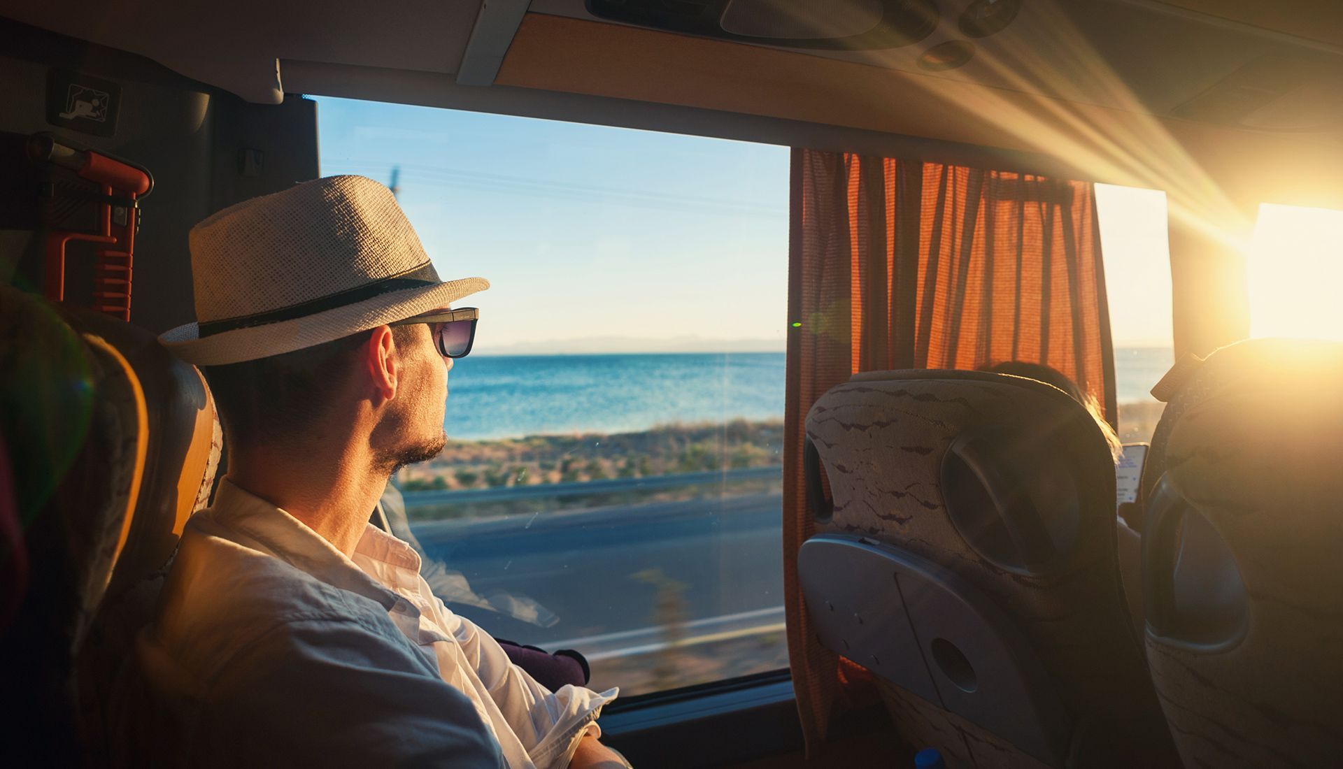 Man in hat and sunglasses gazes out bus window at sea and sunrise.