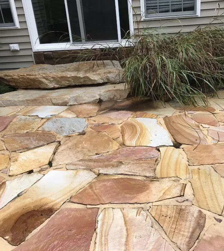 Flagstone patio with steps leading to a sliding glass door. Brown and tan stones. Tall grass grows near the door.