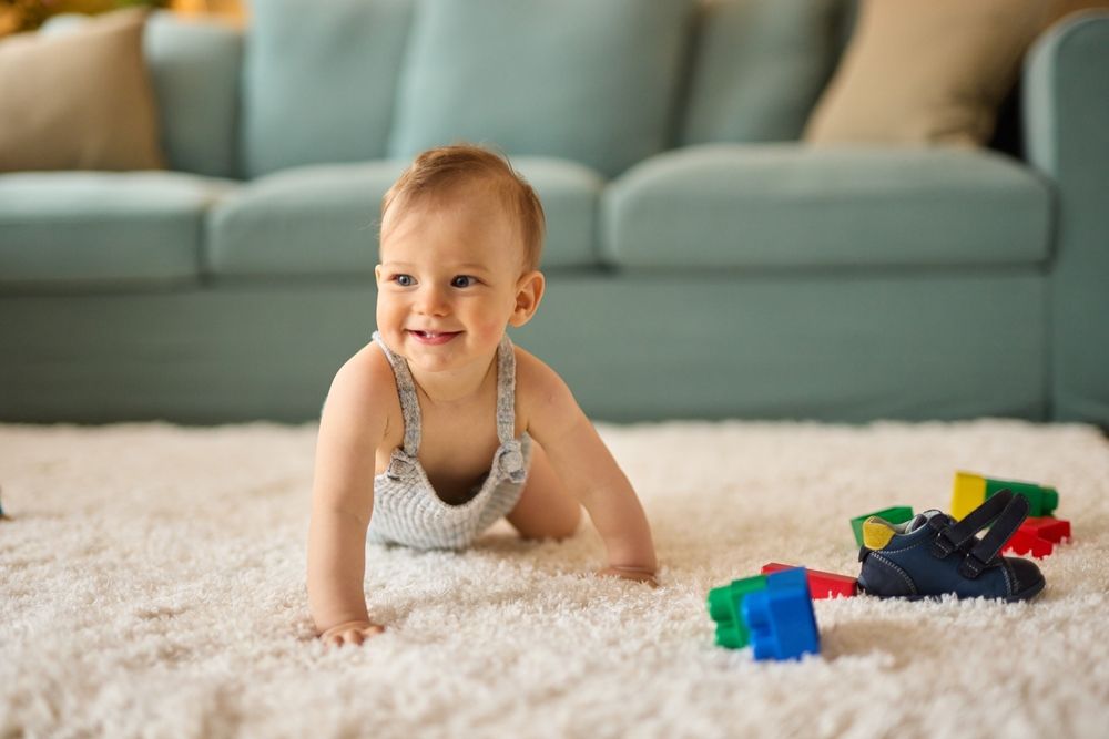 Baby crawling on a white carpet, smiling, with toys and a blue couch in the background.