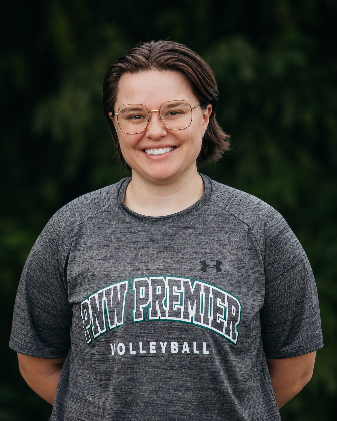 Smiling person in glasses wearing a gray New Paltz Volleyball T-shirt against a dark green background