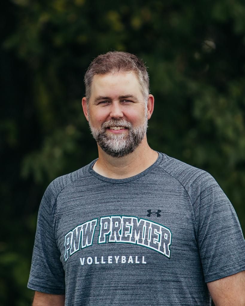 Man in a gray “Pawnee Volleyball” T-shirt standing outdoors against a blurred green background