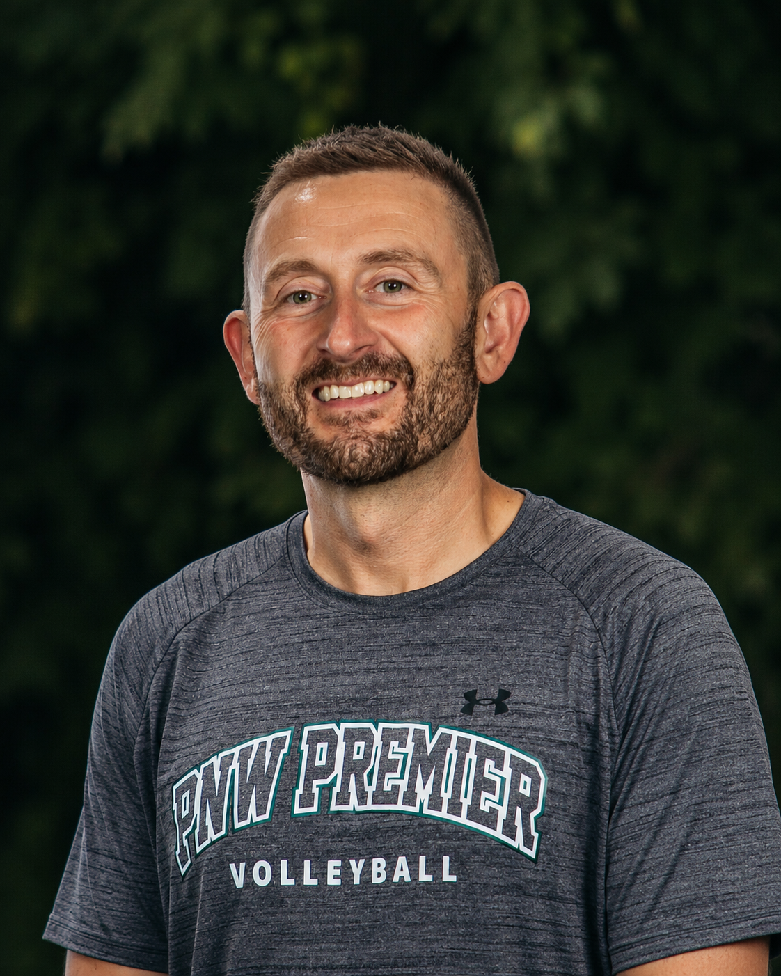 Smiling man in a gray “NW Premier Volleyball” T-shirt against a dark background