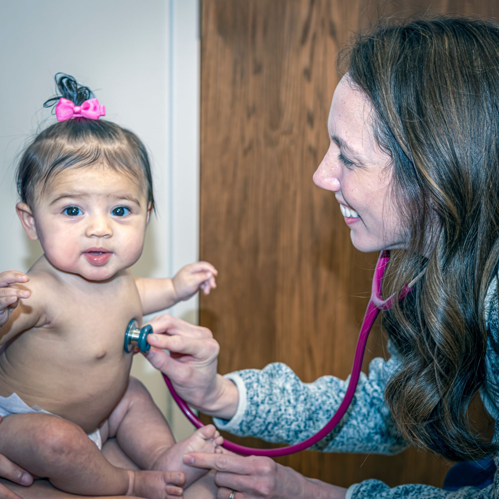 Mom holding a baby in her lap while a pediatrician listens to her heart beat