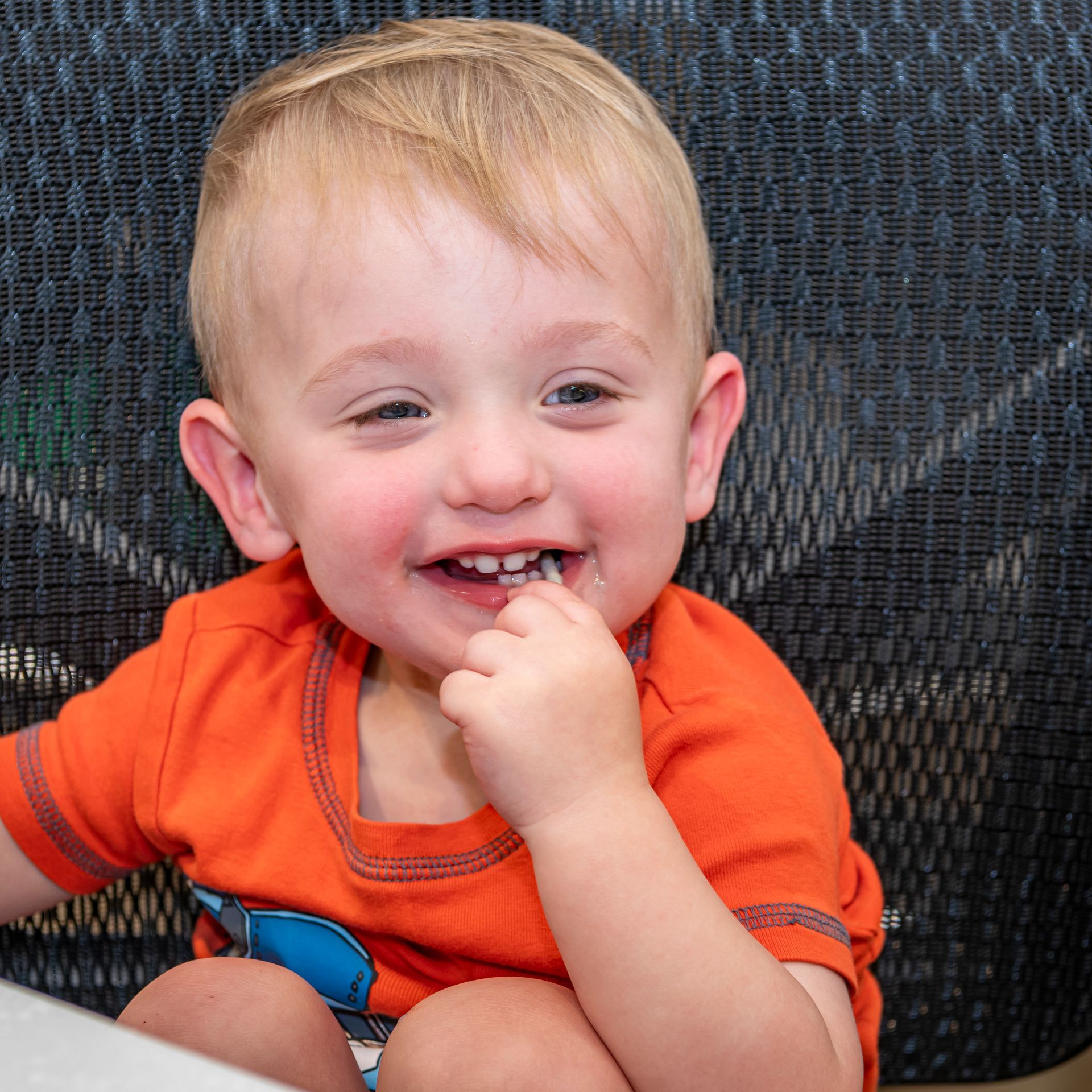Smiling toddler with blonde hair and orange shirt, fingers in mouth.