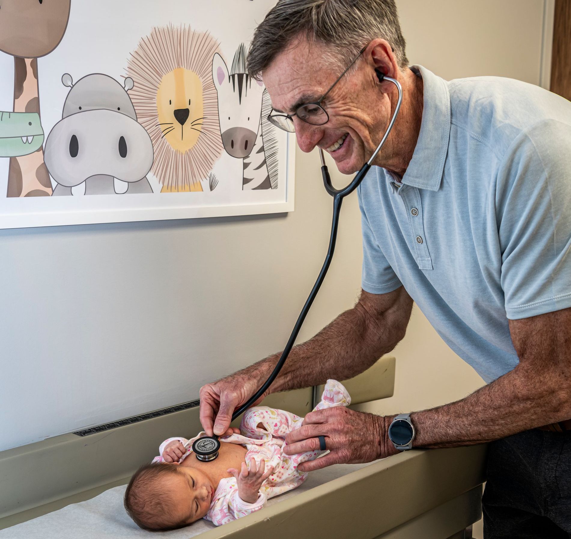 Pediatrician using a stethoscope on a baby, smiling. Neutral examination room with animal artwork.