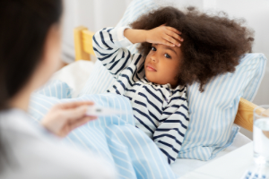 A child lying in bed with a hand on their forehead while a person in the foreground holds a digital thermometer.