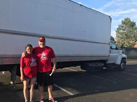 Two people in red shirts stand in front of a large white truck in a parking lot.
