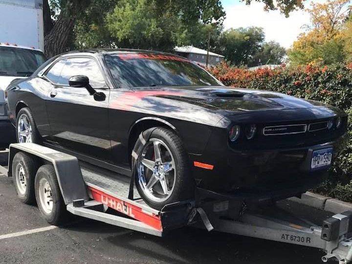 Black Dodge Challenger on a U-Haul trailer, in front of a building, under sunlight.