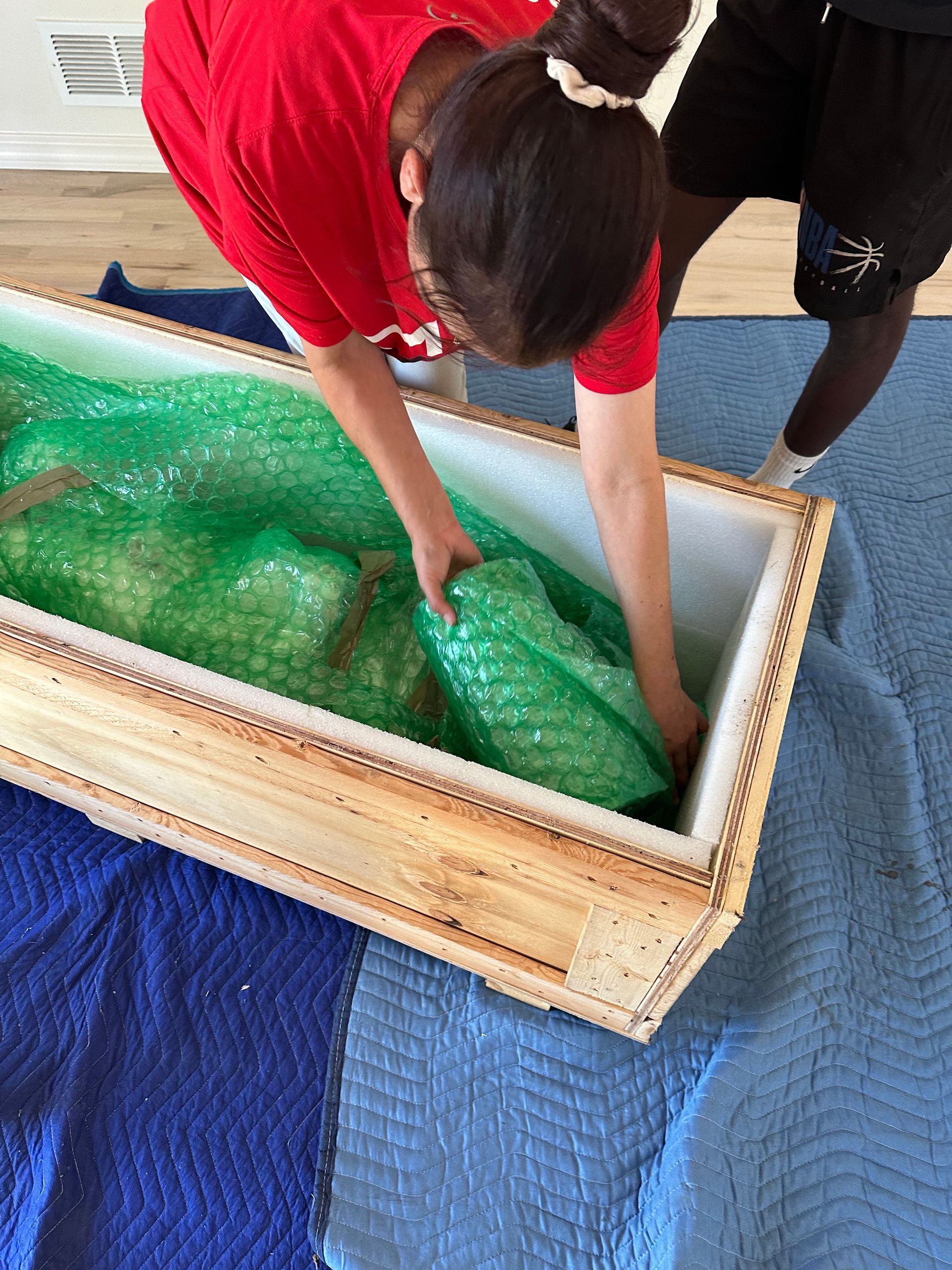 People packing an object wrapped in green bubble wrap into a wooden crate on blue blankets.