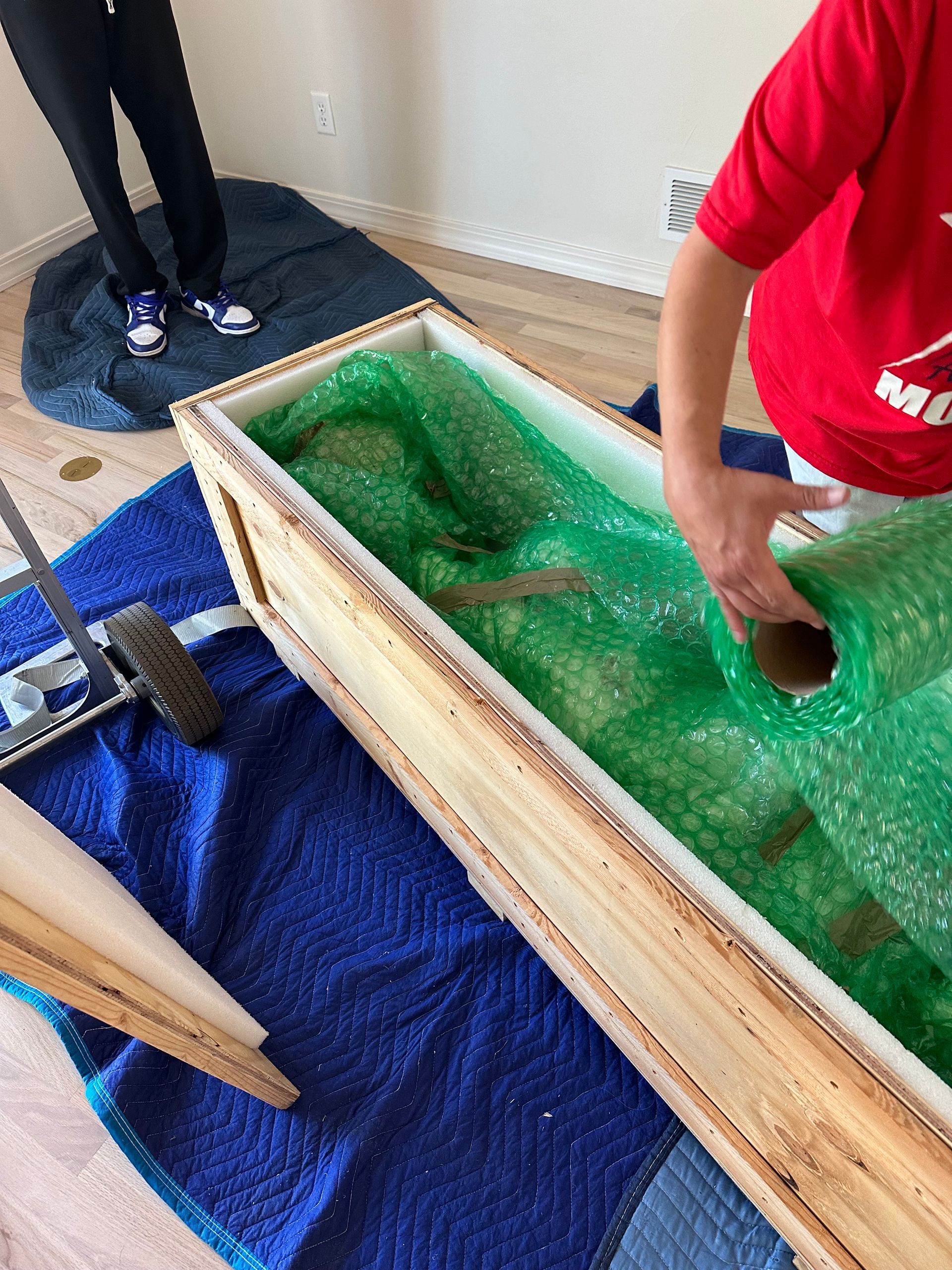 Person packing objects in a wooden crate with bubble wrap. Blue and green colors.
