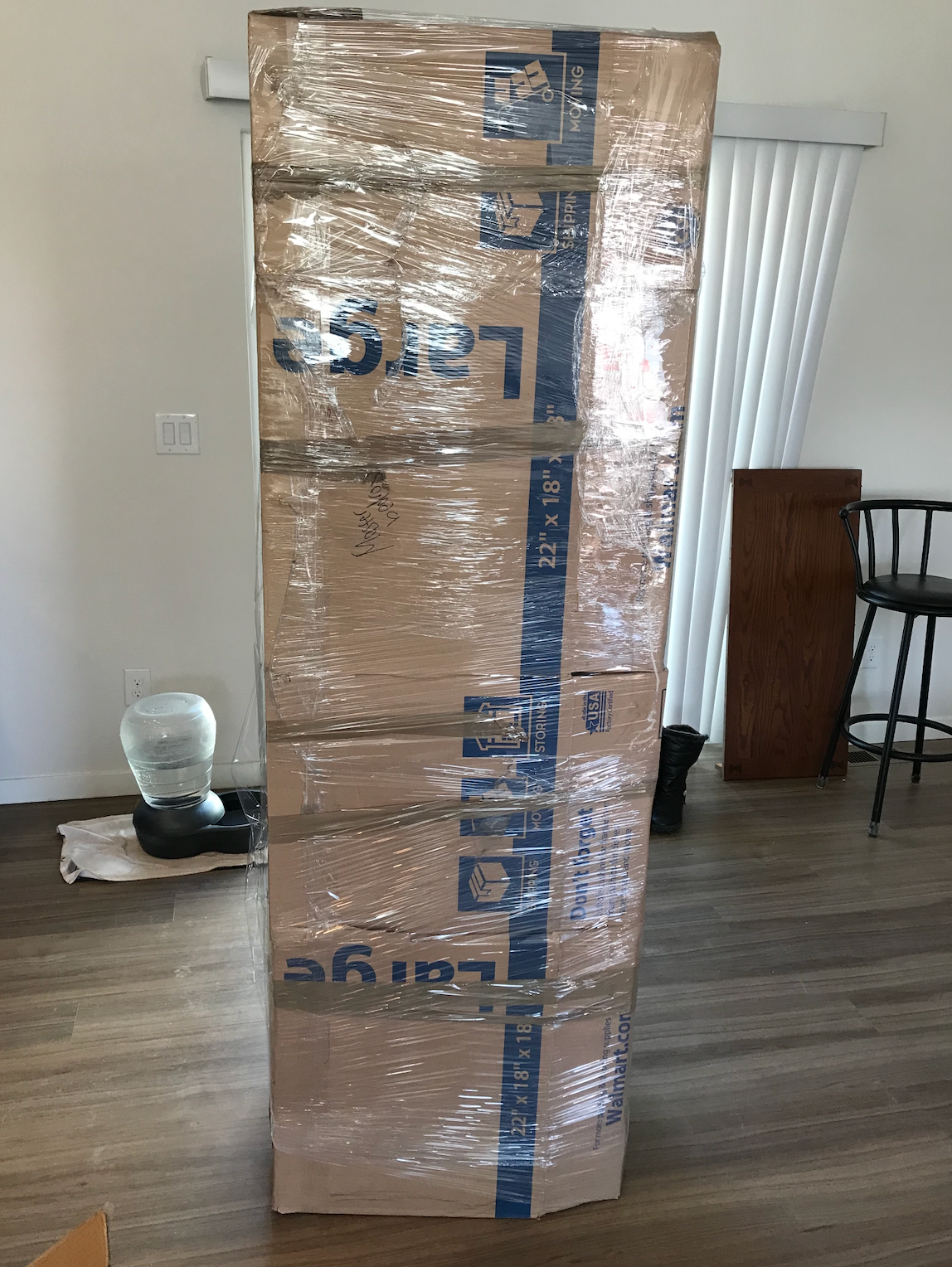 A large cardboard box wrapped in plastic is sitting on a wooden floor in a living room.