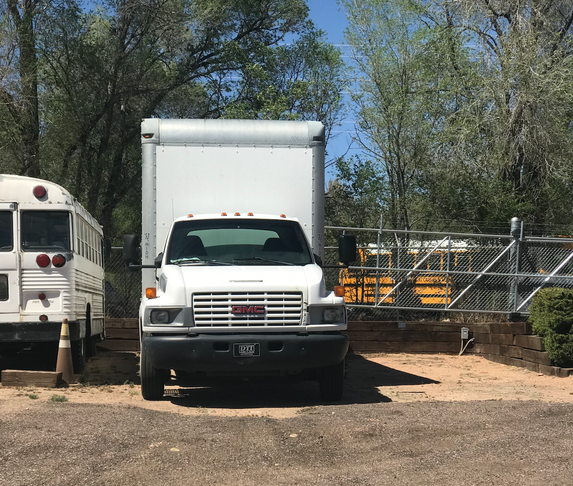 White box truck parked in a gravel lot next to a white school bus and a chain link fence.