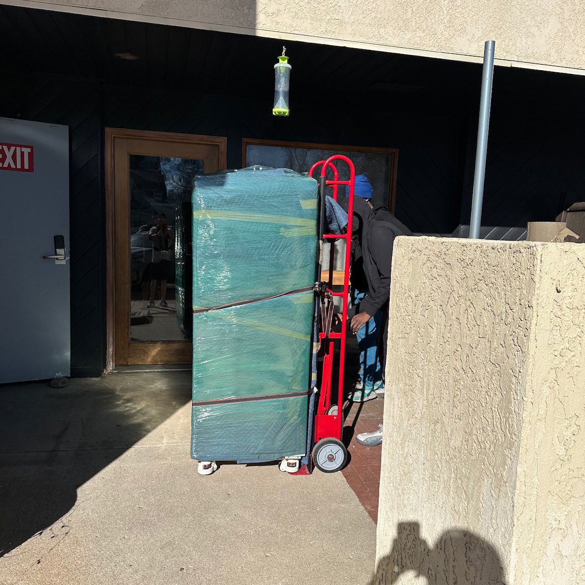 A large, wrapped item is on a dolly being moved by a person near a building entrance with an exit sign.
