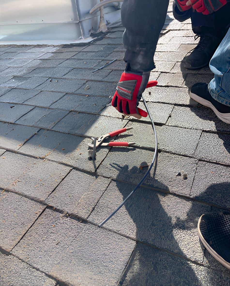 Person on a roof using pliers and hammer to work on shingles. Red gloves are visible.
