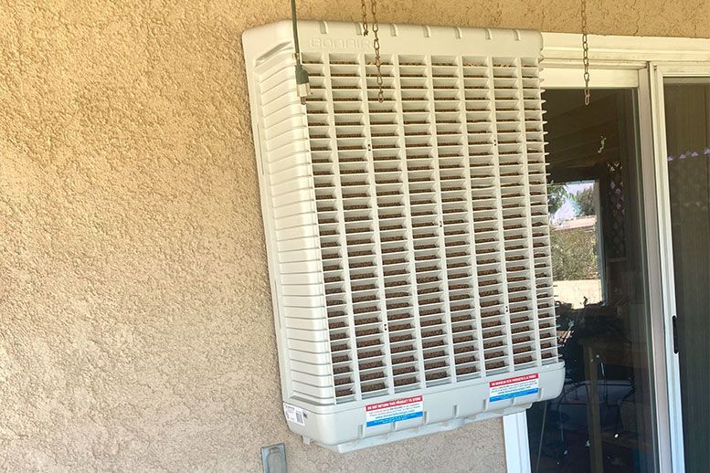 Evaporative cooler, beige with horizontal vents, mounted on a tan stucco wall next to a sliding glass door.