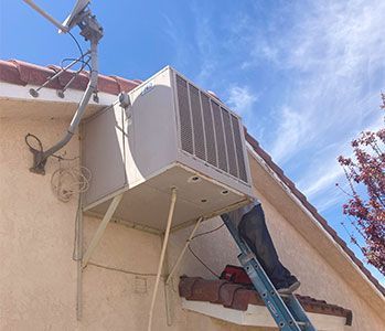 Air conditioning unit mounted on a bracket, being worked on by a person on a ladder outdoors.