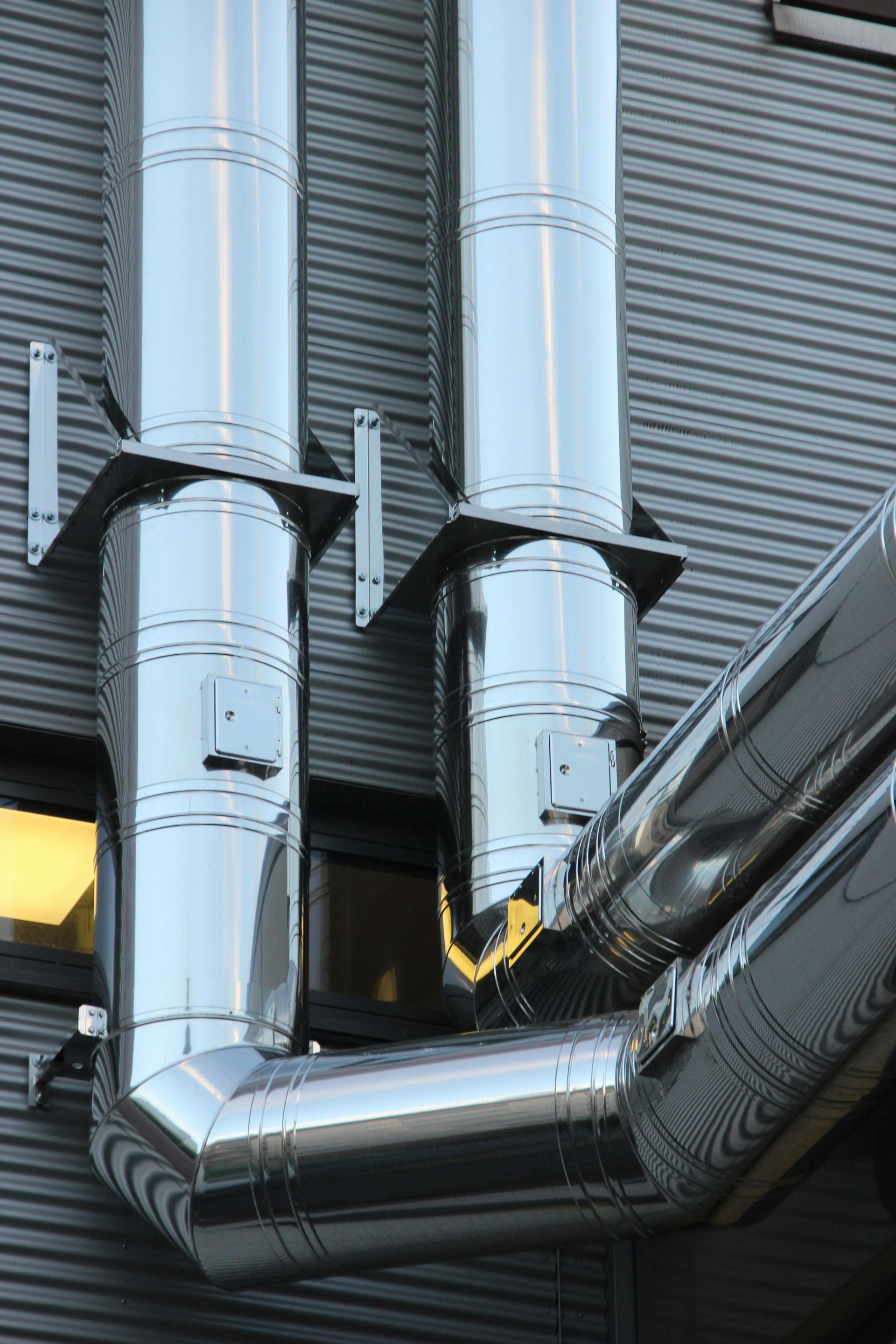 Silver metal pipes attached to building's gray exterior, angled and bent with supports.