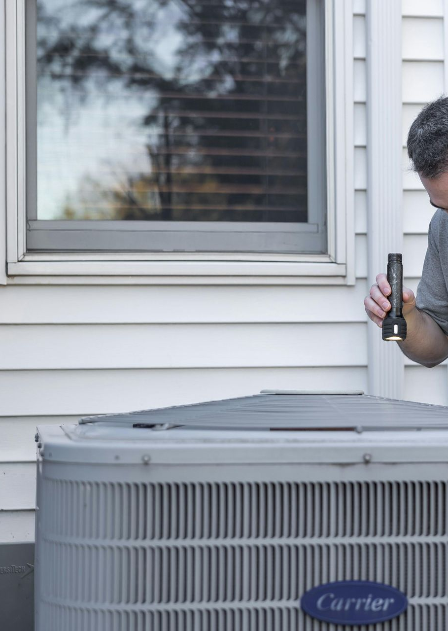 Man checks an air conditioner unit with a tool outside a house, near a window.