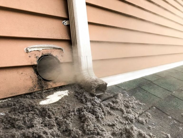 Exterior dryer vent with visible lint and debris, next to a downspout and siding.