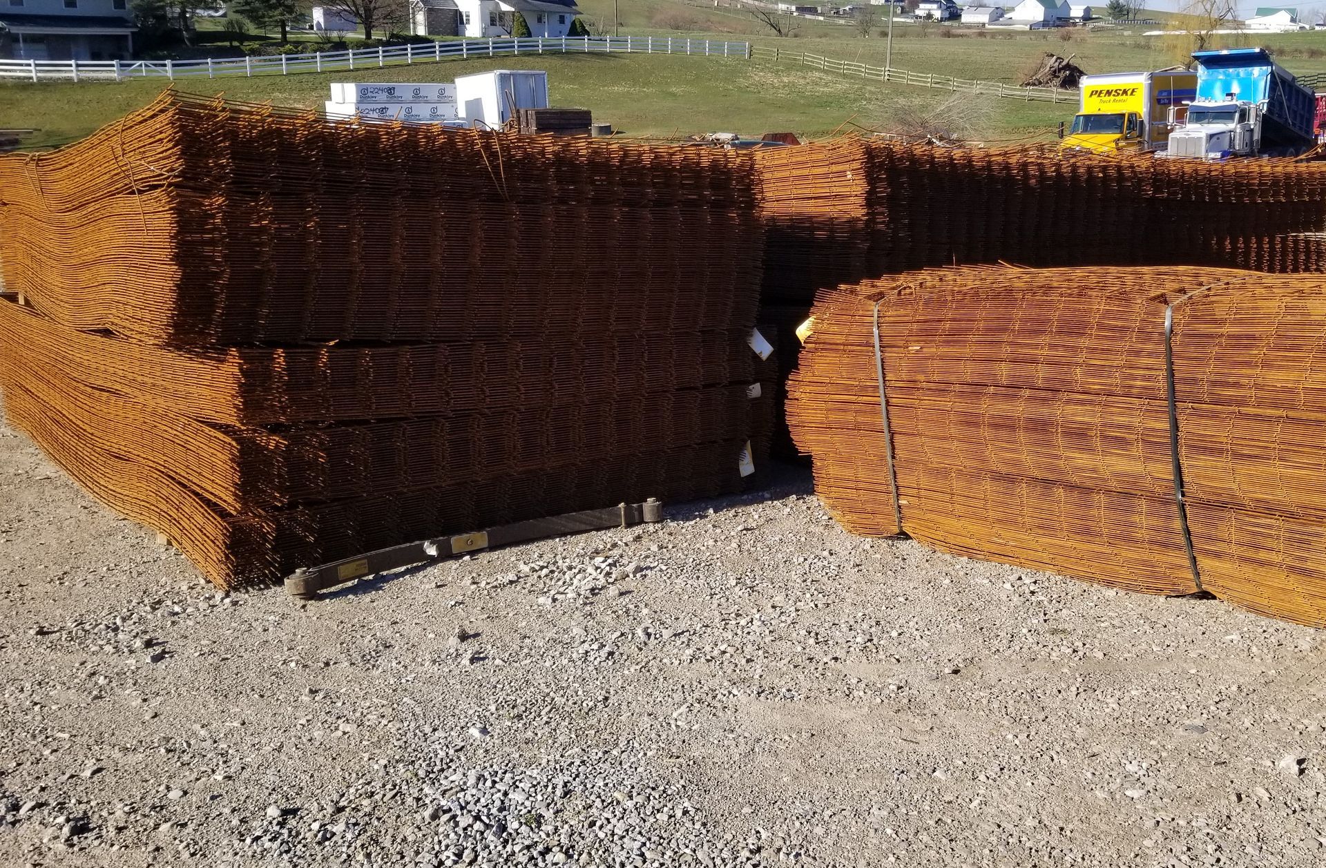 Stacks of rusty rebar mesh on gravel, construction site in the background.