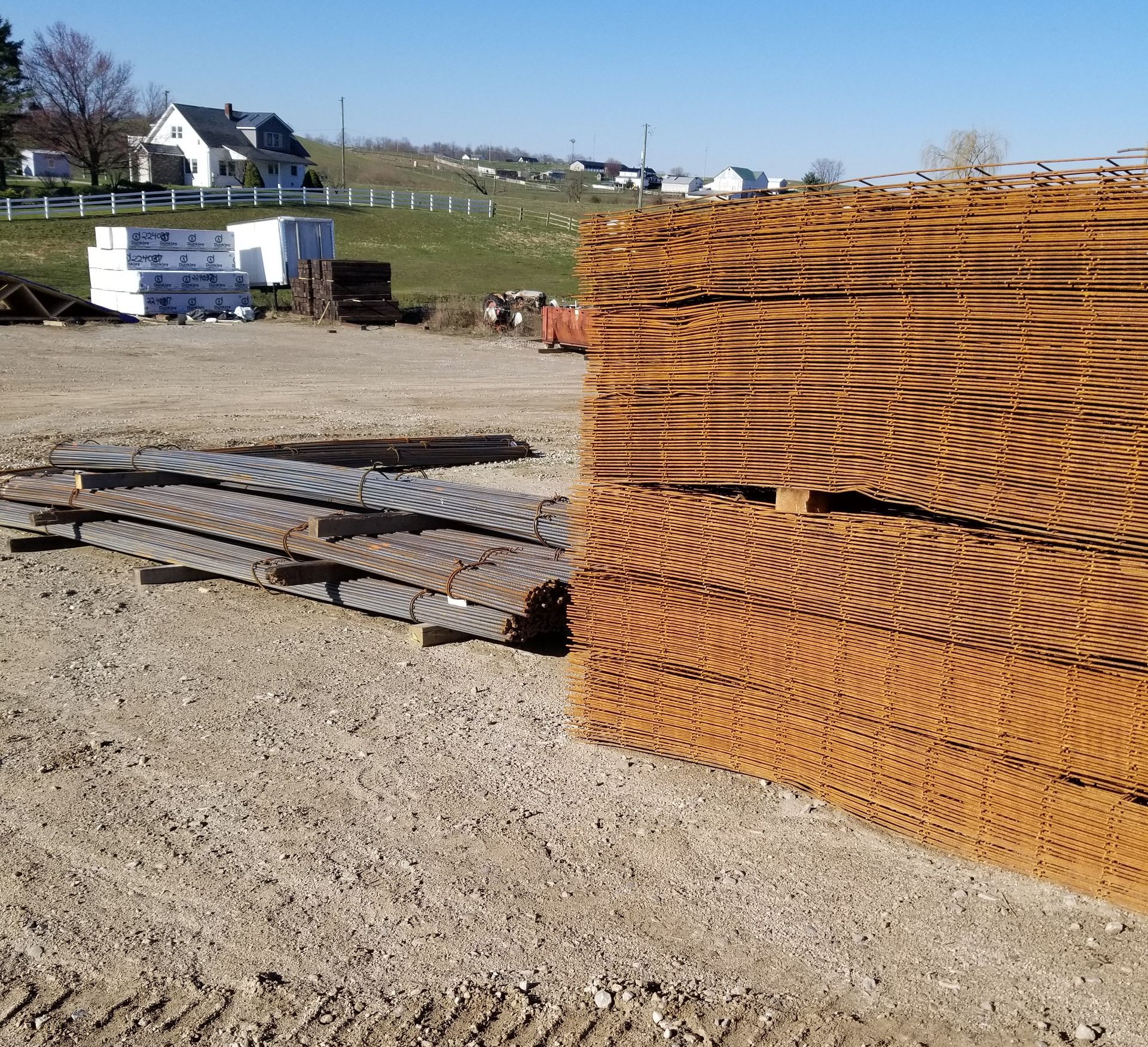 Stacks of rusty metal mesh on a gravel lot; houses and a grassy hill in the background under a blue sky.