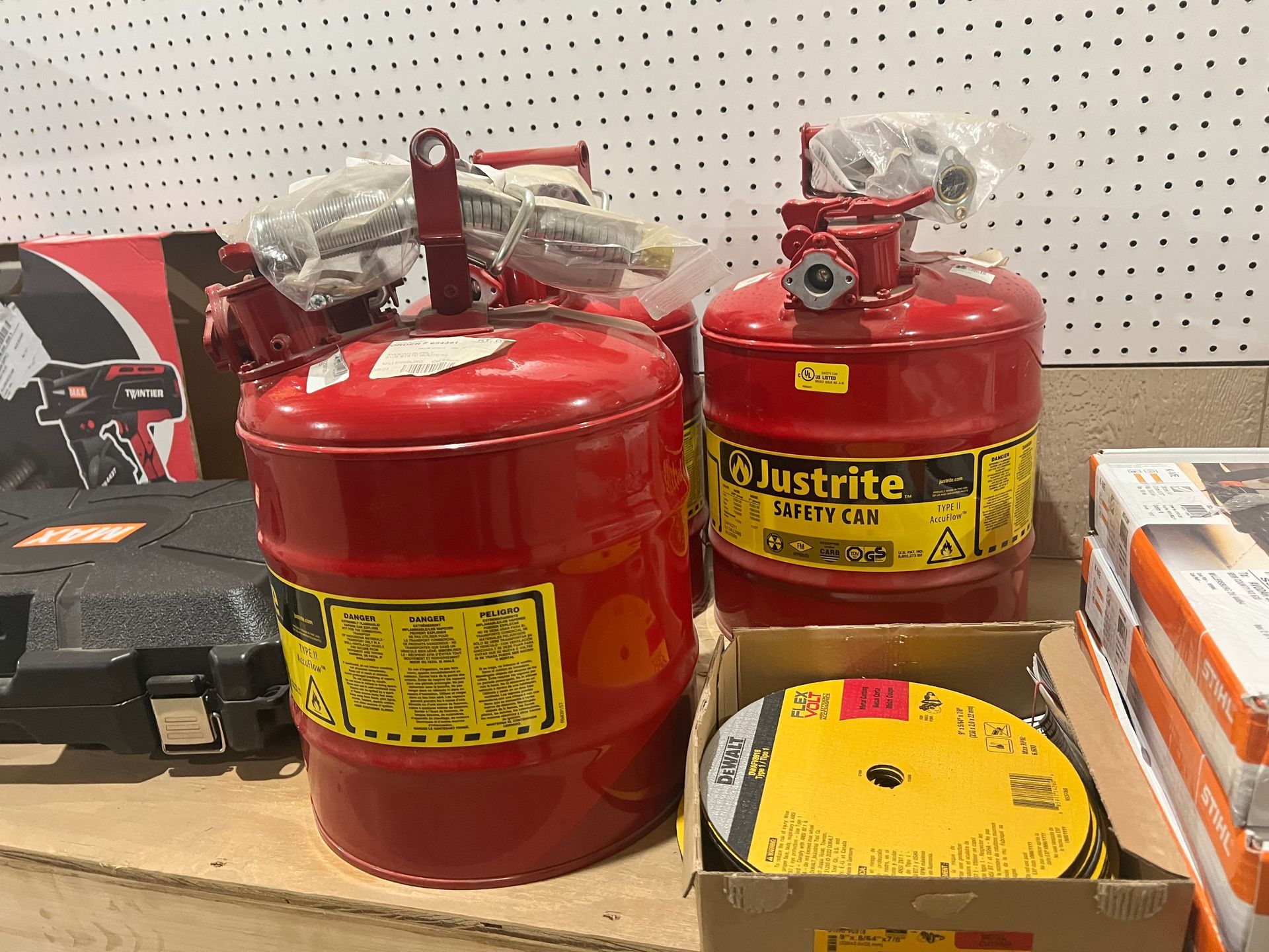 Two red Justrite safety cans with hoses and a yellow label, sitting on a shelf with other items.