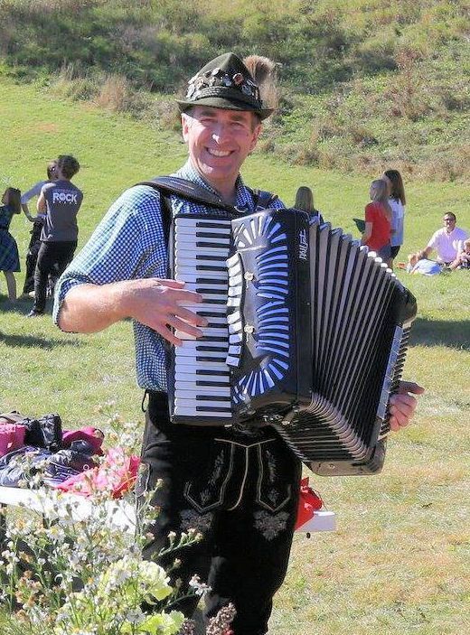 Man playing accordion outdoors, wearing traditional Bavarian attire.
