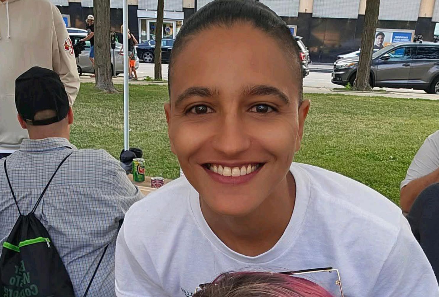 Person smiling, outdoors. Wearing white t-shirt, short dark hair. Background includes people, cars, and buildings.