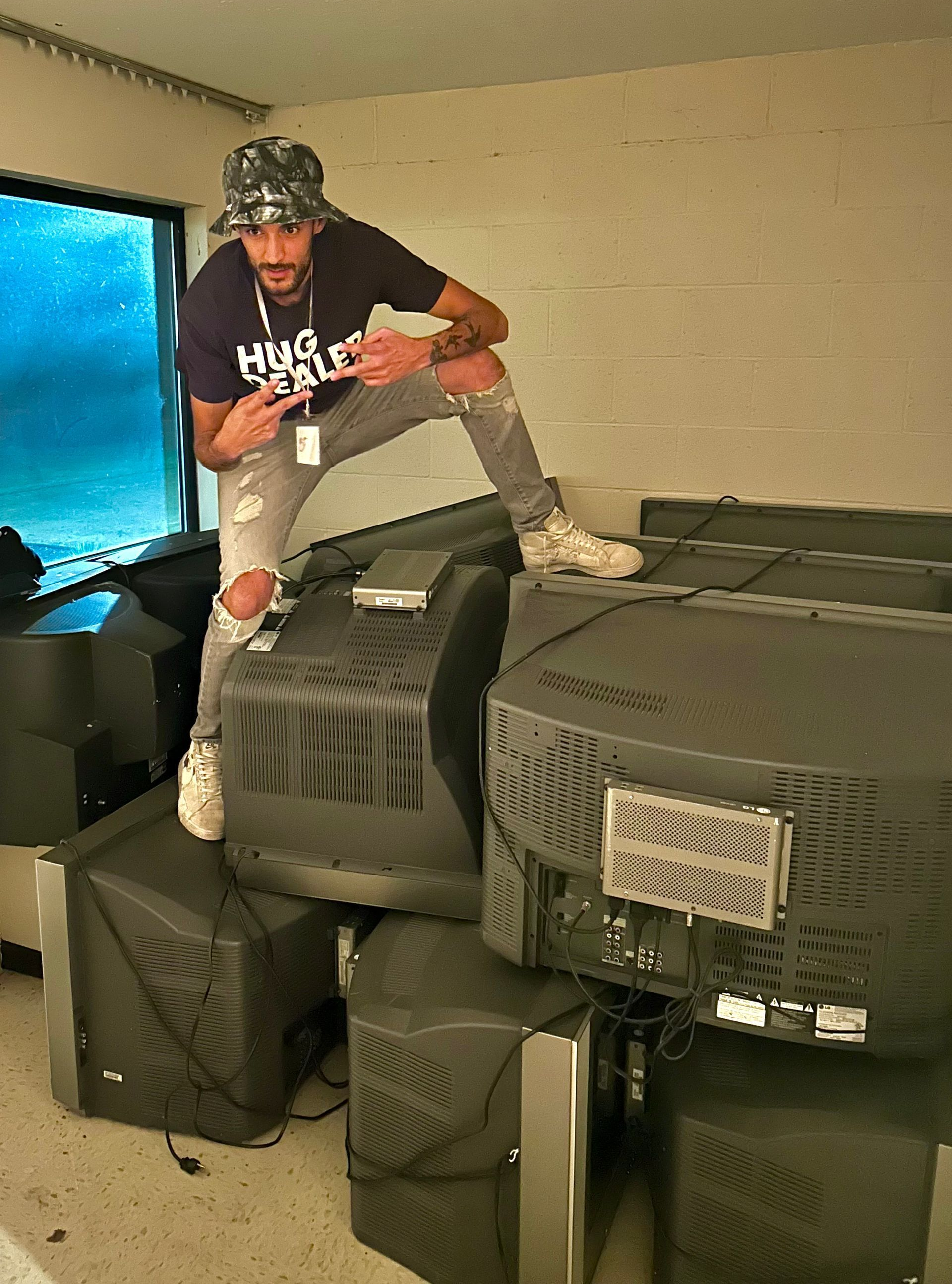 Man on pile of old TVs, wearing hat, ripped jeans, and a t-shirt, pointing.