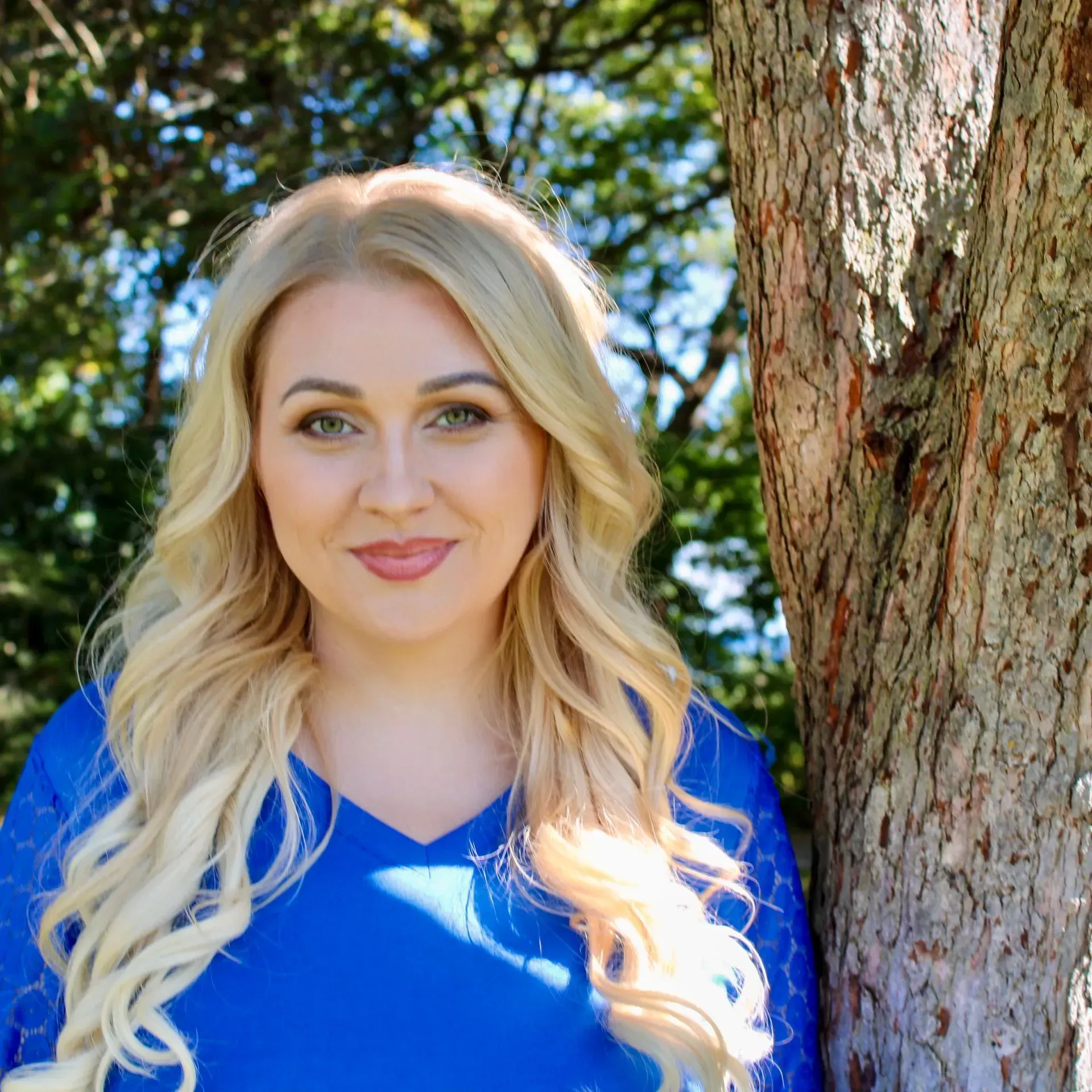 Blond woman in blue top smiles near tree in outdoor setting.