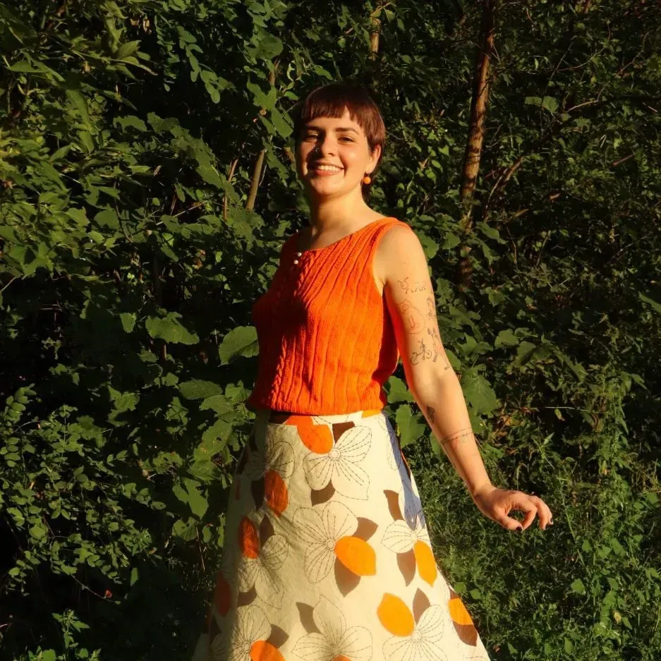 Woman smiling, wearing orange top and floral skirt, standing in front of green foliage.