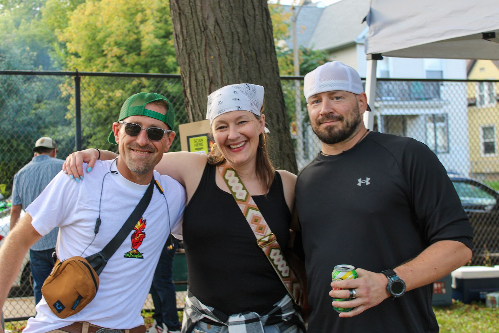 Three people smiling, posing together outdoors by a tree. One wears a green hat, one a bandana, one a white cap.