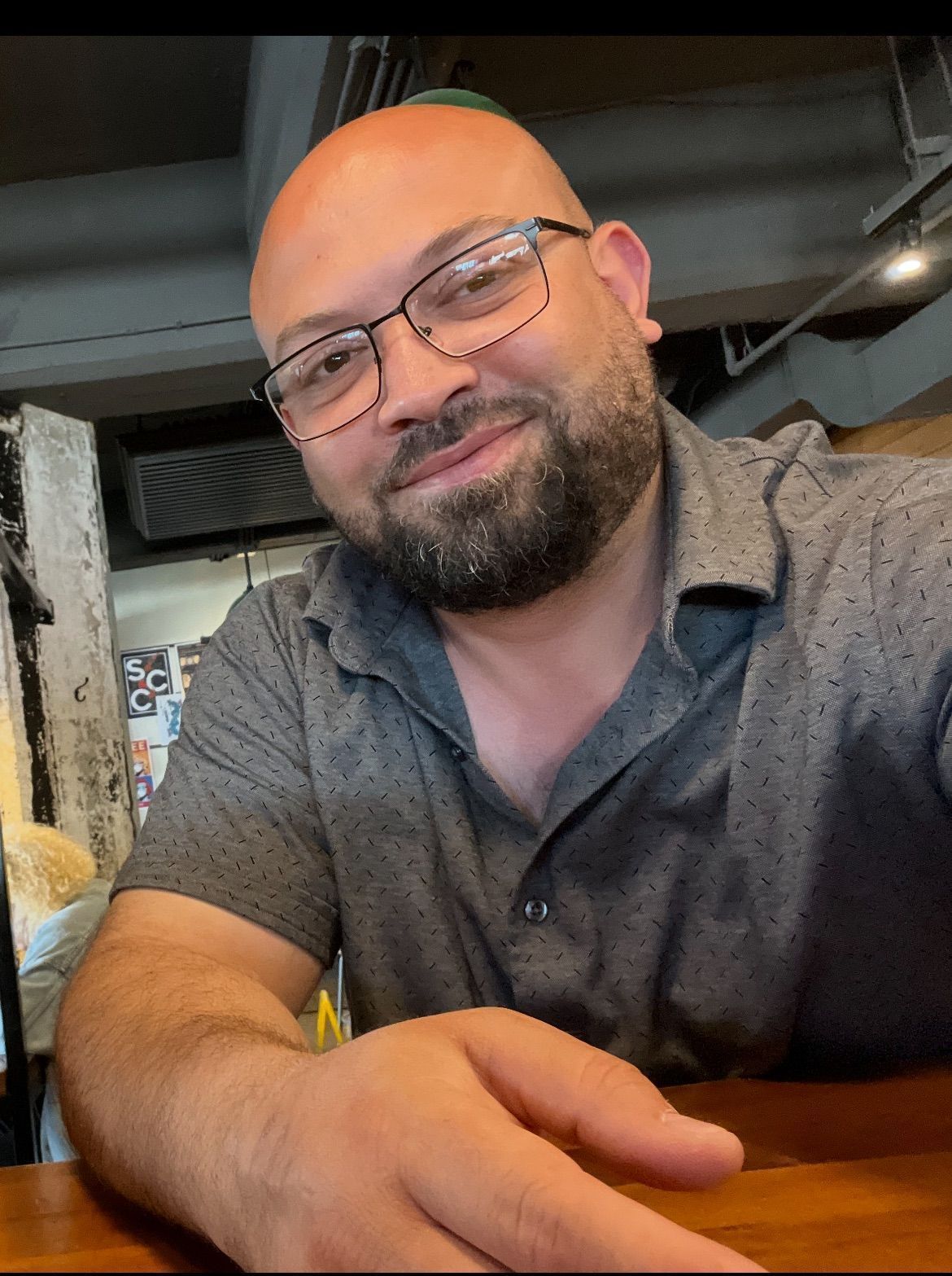 Bald man with glasses, a beard, and a gray patterned shirt, smiling. Indoors, seated at a table.