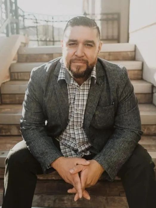 Man sitting on outdoor steps, wearing a patterned shirt and gray blazer, with hands clasped.