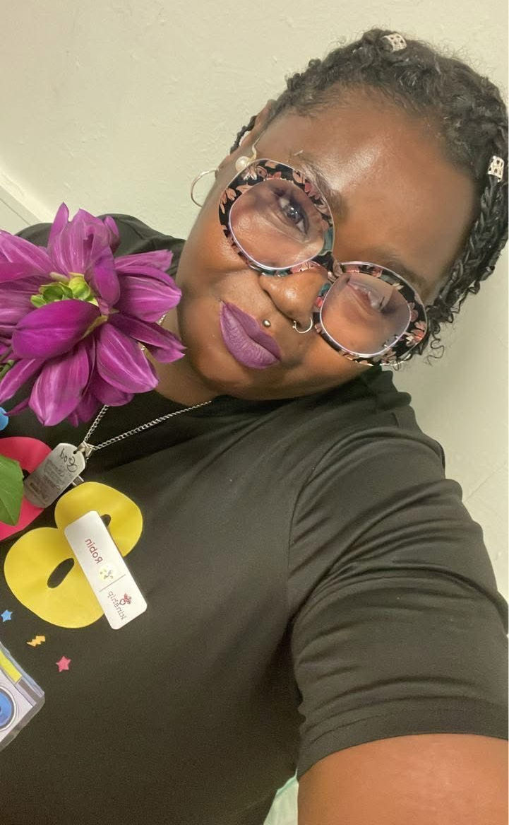 Woman holding purple flower, wearing glasses and black shirt.