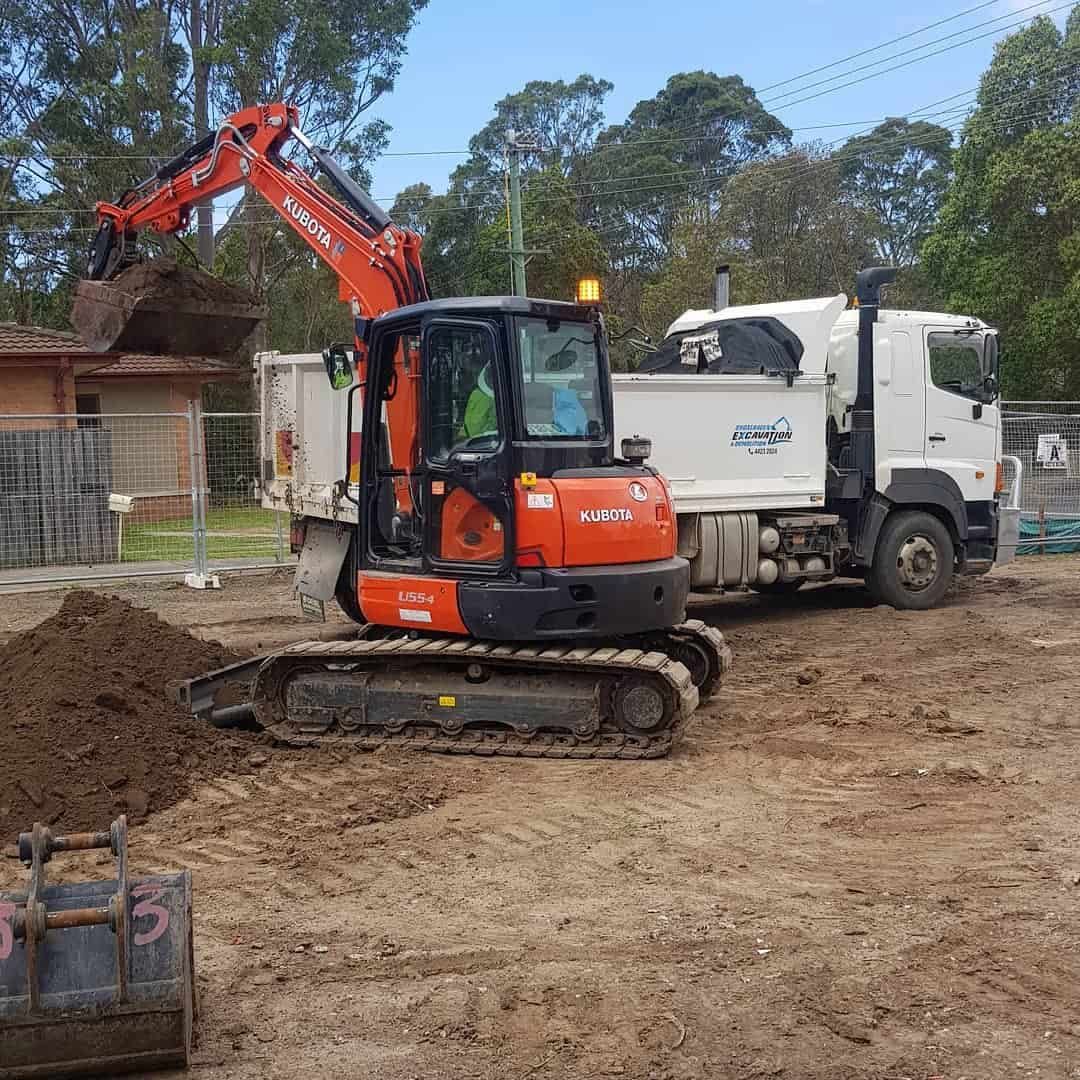 An Orange Excavator Loads Dirt Into a White Dump Truck at a Construction Site — Dirt Werx in Nowra, NSW