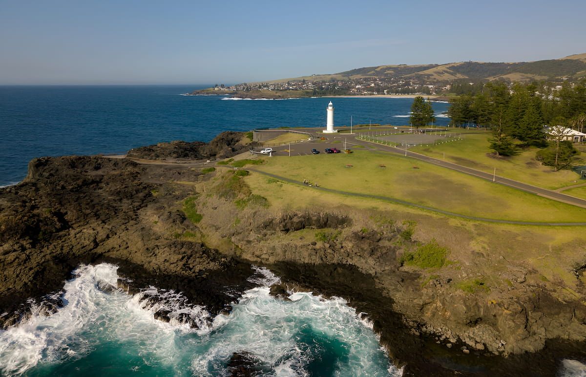 Lighthouse on a Coastal Cliff With Waves Crashing Below and a Distant Town — Dirt Werx in Kiama, NSW