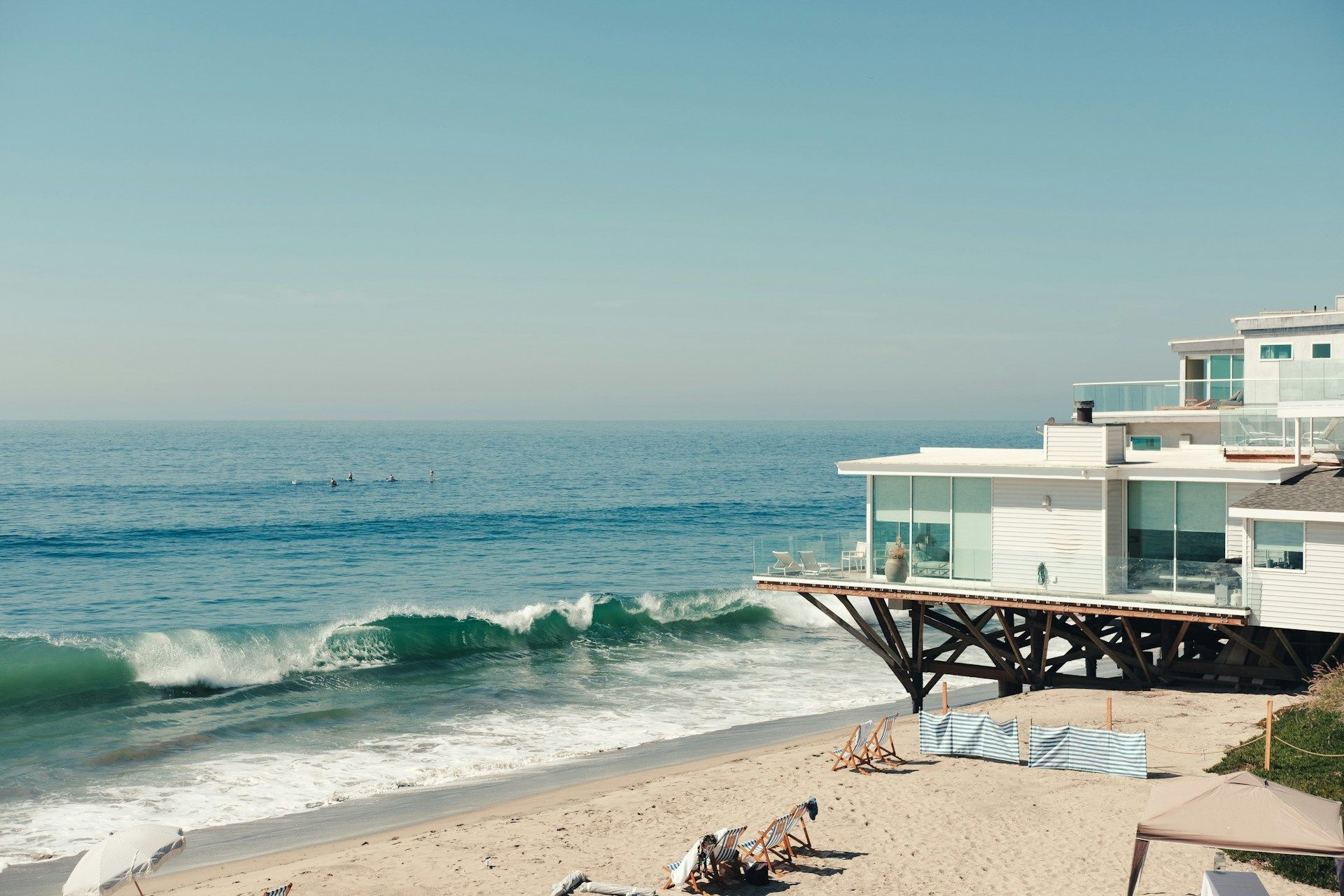 Oceanfront white house with a deck overlooking the sandy beach and crashing waves under a clear blue sky.