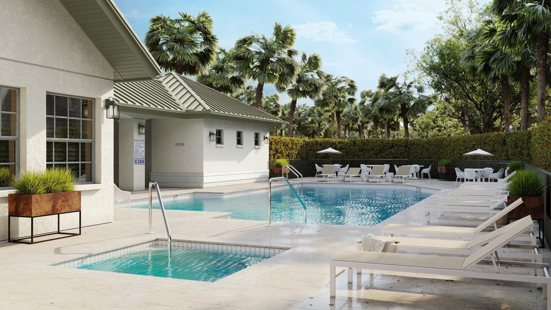 Pool area with white lounge chairs, pool, and small building. Lush green trees.