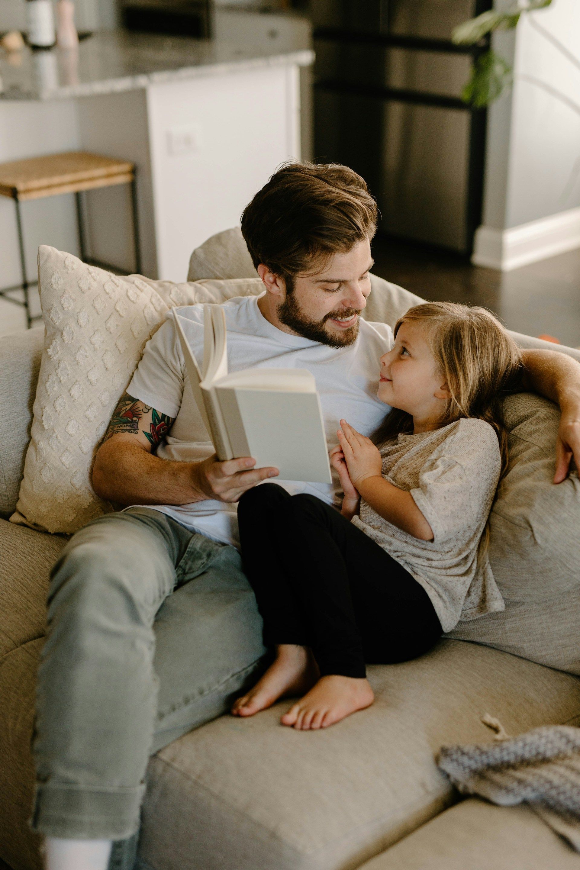 Man reading a book to a child on a beige sofa; both are smiling and looking at each other.
