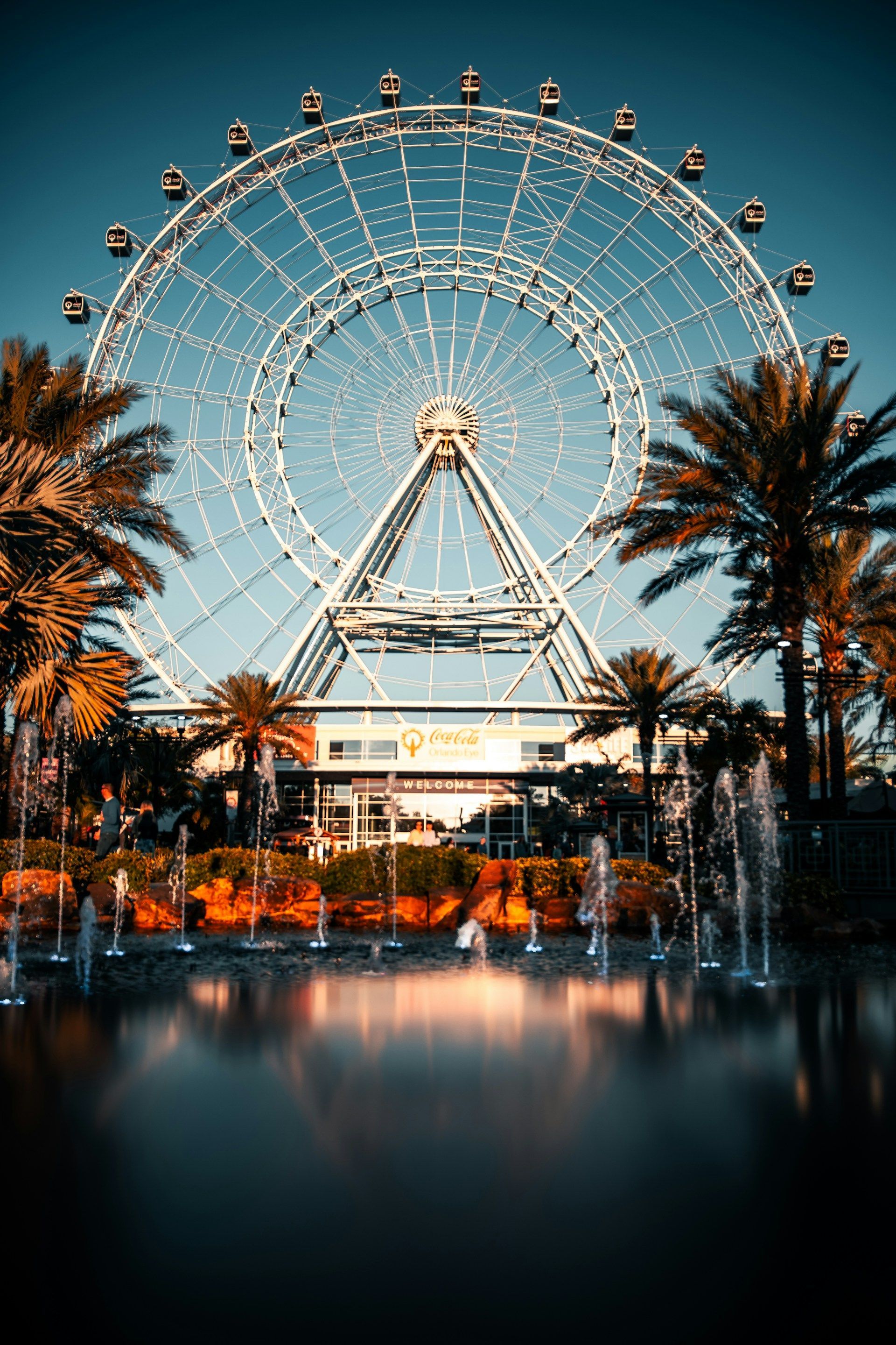 Large Ferris wheel against a blue sky, flanked by palm trees and water fountains.